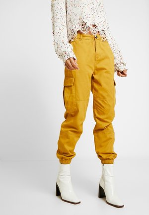 Värikäs neulepusero, jossa leikatut reunat, sinappi-väriset cargohousut joustavilla resoreilla ja valkoiset nilkkurit korolla seisovan henkilön päällä.