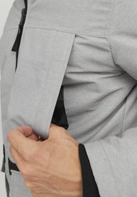 Pilkas švarkas su tekstūruotu paviršiumi, turintis šoninį užtrauktuką ir juodą vidinį pamušalą. Pabrėžtas kontrastuojančiais juodais akcentais ant rankovių.