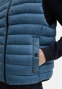 Blauwe puffer vest met gequilte textuur, ritssluiting en zijzakken met een knoop. Draag over een donker t-shirt met lange mouwen.