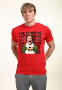 Červené bavlněné tričko s grafikou postavy oblečené v zeleném s bílým kožíškem, obklopené opakujícím se textem: "SANTA PŘICHÁZÍ."