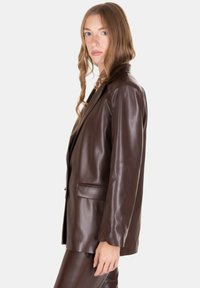 Una giovane donna con lunghi capelli ondulati indossa un blazer in pelle marrone scuro e pantaloni coordinati, in piedi di lato contro uno sfondo neutro.