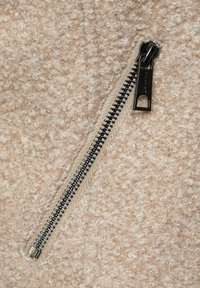 Béžová teddy vlněná bunda s texturovaným povrchem. Obsahuje černý kovový zip s očkem na táhnutí a hnědý látkový lem.