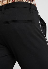 Schwarze Hosen mit schmaler Passform, strukturiertem Stoff, einer einzelnen Gesäßtasche und kontrastierenden weißen Seitenstreifen entlang der Beine.