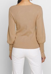 Beige gerippter Pullover mit weitem Halsausschnitt und puffigen langen Ärmeln, kombiniert mit weißen Jeans; der Stoff wirkt weich und hat ein tailliertes Design.