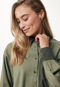 Camicia verde oliva a vestibilità ampia con chiusura a bottoni frontale, maniche lunghe e polsini arricciati. Tessuto morbido e testurizzato con una leggera lucentezza.