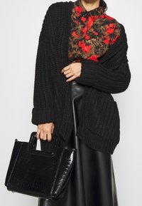 Cardigan de punto grueso negro, camisa con estampado floral en marrón y rojo, y un bolso brillante de textura de cocodrilo negro con asas cortas.