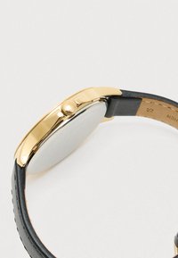 Rund urskive med et gold-tone metalhus, sort læderrem og tekstureret krone. Urets ansigt er glat med et minimalistisk design.