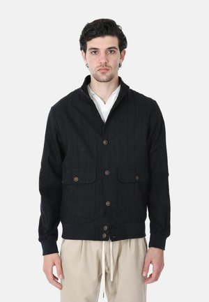 Giovane uomo con capelli scuri e ricci indossa una giacca nera con bottoni sopra una camicia bianca e pantaloni beige con coulisse, in piedi davanti a uno sfondo semplice.