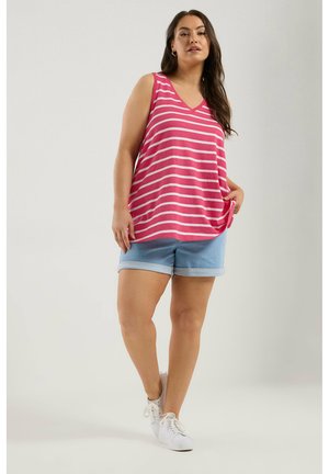 Kobieta ubrana w różowo-biały top bez rękawów w paski, jasnoniebieskie szorty z denimu i białe trampki, stojąca z jedną ręką w kieszeni.