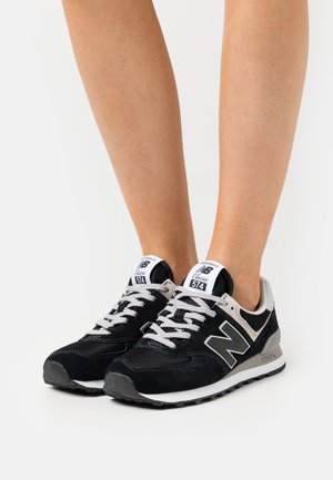 Jambes portant des baskets New Balance 574 Classic noires et blanches avec des lacets blancs, debout sur une surface blanche.