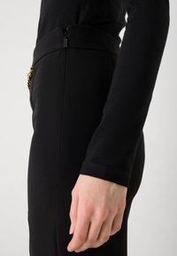 Čierne elastické nohavice s vysokým pásom, obsahujúce malý bočný zips a reťazový detail na páse. Hladká textúra.