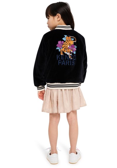 Veste en velours noir ornée d'un tigre brodé et d'un motif floral, avec le texte "KENZO PARIS", des poignets et un ourlet rayés. Portée sur une jupe rose clair.