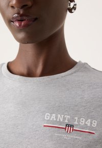 Siva bombažna majica s krožnim izrezom, na kateri je bel napis "GANT 1949" in zaščitni znak v rdeči, beli in modri barvi.