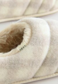 Fuzzy cream and beige slippers with a plush interior and stitched stripes. Label on the side reads "NEXT." Soft texture and cozy design.