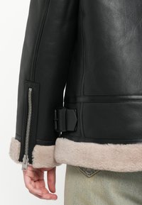 Zwarte leren jas met schapenvachtaccent, met een zijrits en een gespdetail in de taille. Zachte textuur, gestructureerd ontwerp.