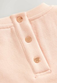 Detalle de un suéter rosa claro que presenta un cuello acanalado, tres botones de presión de metal y una tela suave y texturizada.