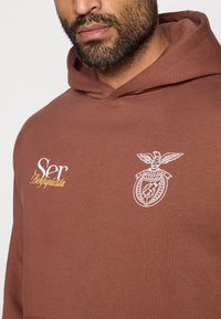 Brązowy bawełniany hoodie z białym logo godła orła i białym napisem "Ser Benfiquista" na klatce piersiowej. Przytulny, luźny krój z kapturem.