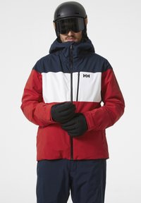 Veste de ski colorée en blocs rouge, blanc et bleu marine avec un col haut, gants noirs et casque de ski. Comprend une fermeture éclair à l'avant et un détail du logo de la marque.