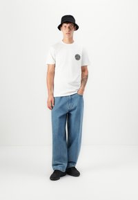 Hvit bomull t-skjorte med rund svart logo på brystet, kombinert med løse blå denimjeans og svart bøttehatt, fullført med svarte sneakers.
