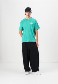 T-shirt verde oversized com um pequeno logotipo branco, combinada com calças largas pretas. Modelo usa um boné preto e tênis brancos.