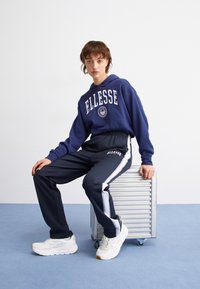 Jūras zila kapuce ar baltu "ELLESSE" logo, saskaņota ar jūras zila sporta biksēm ar sānu līnijām un baltām kedām ar pelēkiem akcentiem. Sēž uz sudraba konteineru.