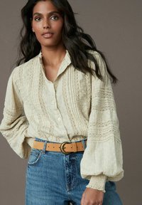Blouse crème avec un motif texturé, dotée d'un devant à boutons et de manches fluides, associée à un jean bleu taille haute et une ceinture marron.