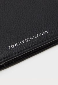 Portefeuille en cuir noir à finition texturée. Présente un texte en relief blanc "TOMMY HILFIGER" ainsi qu'un logo. Forme compacte et rectangulaire.