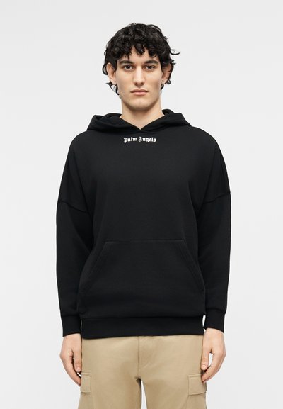 Mees lokkis tumedate juustega, kandes musta Palm Angelsi kapuutsiga pusa ja beeže lahtiste taskutega pükse, seisab heleda ühevärvilise tausta ees.