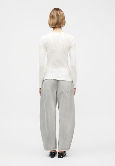 Γυναίκα με κοντά ξανθά μαλλιά που φοράει λευκή μαλακή μπλούζα με μακριά μανίκια, χαλαρό ανοιχτό γκρι τζιν και μαύρα παπούτσια με καρφιά, όρθια στραμμένη προς τα πίσω.