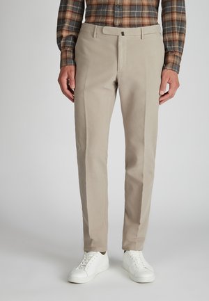 Mann in beigefarbenen Slim-Fit-Hosen, weißen Sneakers und einem langärmligen Hemd in Braun und Grau mit Karomuster, der vor einem grauen Hintergrund steht.