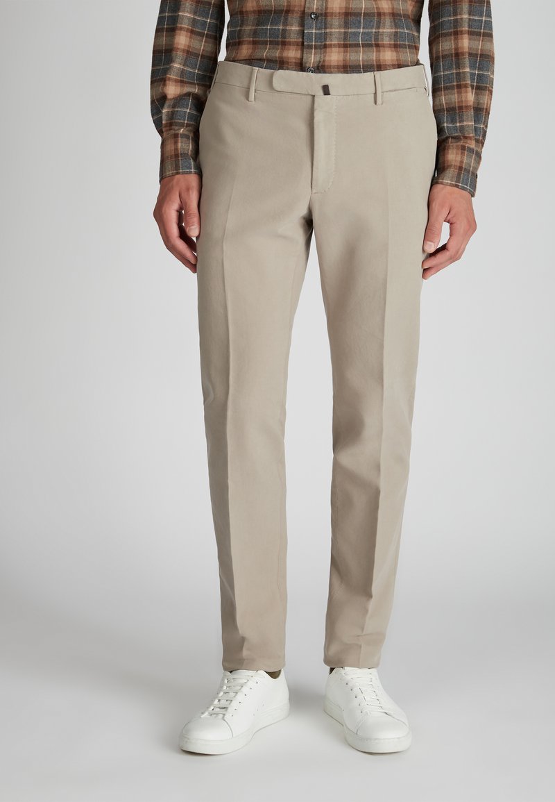 Un uomo che indossa pantaloni beige slim-fit, sneakers bianche e una camicia a maniche lunghe a quadri marrone e grigia, in piedi su uno sfondo grigio.