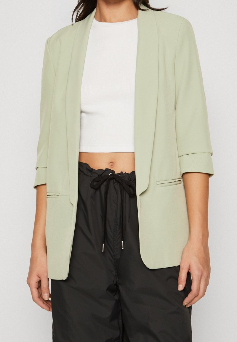 Světlý zelený blejzr se tříčtvrtečními rukávy, otevřený vpředu a s bočními kapsami, kombinovaný s bílým crop topem a černými kalhotami. Hladká textura.