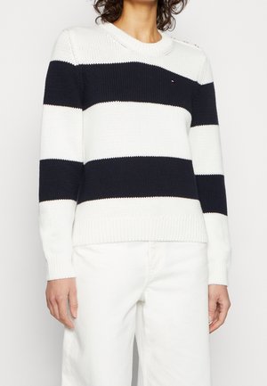 Osoba ubrana w biały sweter z dwoma szerokimi poziomymi granatowymi paskami oraz białe spodnie, widziana od ramion do bioder na jednolitym tle.