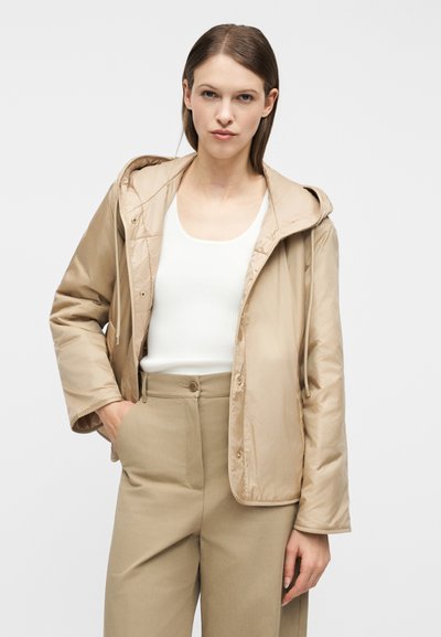 Γυναίκα που φοράει μπεζ επενδυμένο μπουφάν με κουκούλα, λευκό ριμπ μπλουζάκι χωρίς μανίκια και μπεζ ψηλόμεσο παντελόνι, στέκεται με το ένα χέρι στην τσέπη.