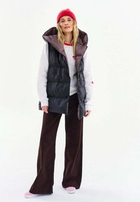 Žena nosi červenú čiapku, bielu mikinu s červenými detailmi, čiernu prešívanú vestu, tmavé široké nohavice a ružové topánky, stojaca na bielom pozadí.