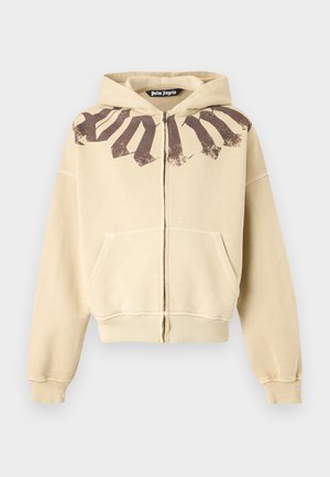 Personne portant un sweat à capuche beige zippé avec un texte marron, un pantalon large sombre et surdimensionné, des baskets rouges et des lunettes de soleil noires, debout devant un fond blanc.