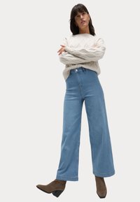 Jean large bleu clair à taille haute, avec fermeture à bouton sur le devant et détails de couture subtils, associé à un pull en tricot torsadé crème.