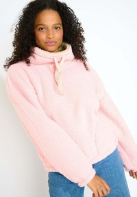 Pullover in pile rosa con colletto alto, tasca frontale e cordino. Tessuto morbido con maniche larghe e orlo aderente. Abbinato a jeans blu.