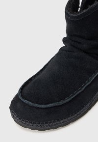 Botín de ante negro con un diseño de parte superior curvada, costuras visibles y superficie texturizada, diseñado para la comodidad y el calor.