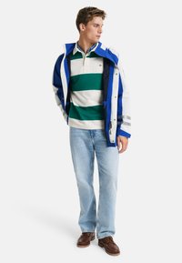 Heren blazer met blauwe en witte strepen, groene en witte gestreepte polo, lichtblauwe jeans en bruin veter schoenen. Kenmerken: zakken en ritsen.