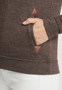 Brun hettegenser med teksturert stoff, med sidelomme og lærtriangelakenter. Mansjettene er tettsittende, og kanten er ribbestrikket.