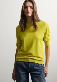 Maglione giallo brillante con colletto e polsini a coste, caratterizzato da una vestibilità rilassata. Abbinato a jeans blu scuro, leggermente usurati.