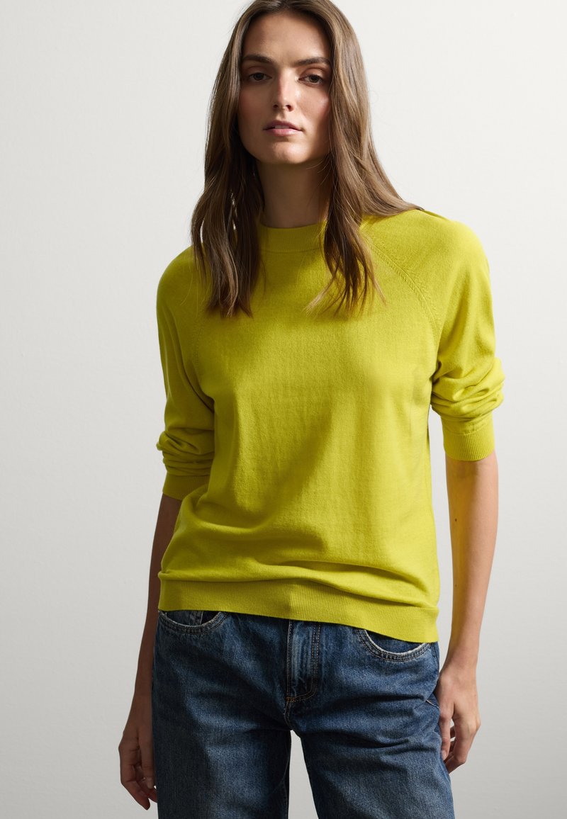 Maglione giallo brillante con colletto e polsini a coste, caratterizzato da una vestibilità rilassata. Abbinato a jeans blu scuro, leggermente usurati.