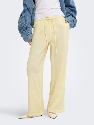 Personne portant un pantalon large à cordon de serrage jaune pâle, des chaussures blanches pointues, un haut blanc et une veste en jean ample avec les mains dans les poches.