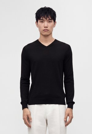 Jeune homme debout, face à l’objectif, portant un pull noir à col en V et un pantalon blanc sur un fond clair uni.