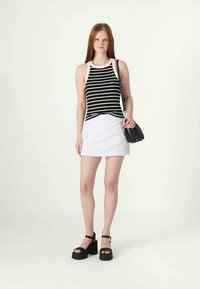 Jovem mulher vestindo uma blusa sem mangas listrada em preto e branco, saia mini branca, sandálias de plataforma pretas e carregando uma mala de ombro preta.