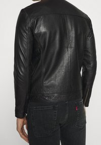 Person i en åtsittande svart läderjacka och mörkgrå denimjeans, visad bakifrån med armarna avslappnat vid sidorna.