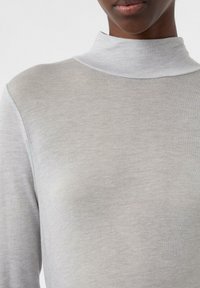 Helles graues Rollkragenshirt mit einer weichen, gerippten Textur, enganliegendem Design und langen Ärmeln, das unter sanfter Beleuchtung einen subtilen Glanz hat.
