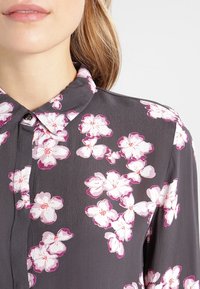 Blouse florale grise en tissu doux avec de grands motifs de fleurs roses et blanches, fermeture par boutons, et col classique.