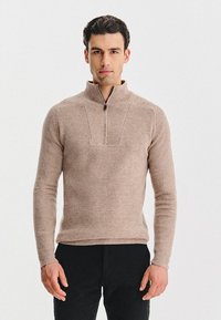Beżowy sweter z dzianiny z kołnierzem na połowie zamka błyskawicznego, prążkowaną strukturą i dopasowanym krojem. Nosi czarne spodnie; model stoi na białym tle.
